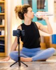 Mujer tranquila con el pelo rizado en ropa deportiva sentada en el suelo y bebiendo botella de agua durante el descanso en la sesión de yoga en línea en el estudio - foto de stock