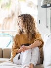 Junge blonde Frau mit lockigem Haar in lässiger Kleidung sitzt auf einem bequemen Sofa, während sie in einem hellen Raum gesunde Nahrung zu sich nimmt — Stockfoto
