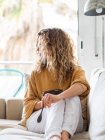 Jeune femme blonde aux cheveux bouclés en vêtements décontractés assis sur un canapé confortable tout en mangeant des aliments sains dans la pièce lumineuse — Photo de stock