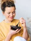 Joven rubia alegre mujer en ropa casual comiendo comida saludable del tazón de coco natural en la sala de luz - foto de stock