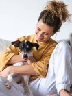Cheerful woman with curly hair an casual clothes sitting with obedient Ratonero Bodeguero Andaluz dog on sofa in light apartment — Stock Photo