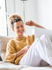 Giovane donna positiva con i capelli ricci in abiti casual seduta su comodo divano e navigazione netbook in appartamento leggero durante il giorno — Foto stock