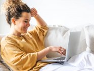 Mujer joven positiva con el pelo rizado en ropa casual sentado en cómodo sofá y netbook de navegación en el apartamento de luz en el día - foto de stock