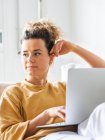 Positive junge Frau mit lockigem Haar in lässiger Kleidung sitzt auf bequemen Sofa und blättert Netbook in hellen Wohnung tagsüber — Stockfoto