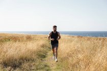 Muscular masculino corredor correndo ao longo do caminho no prado durante o treinamento no fundo do mar no verão — Fotografia de Stock