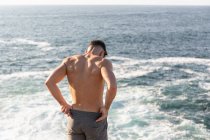 Вид сзади на мускулистого спортсмена с обнаженным туловищем, стоящим на пляже и наслаждающимся закатом после тренировки летом — стоковое фото