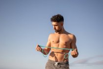 Muscular fisiculturista masculino com tronco nu em pé na praia e fazendo exercícios com banda de resistência durante o treino no verão — Fotografia de Stock