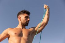 Muskelkräftiger männlicher Bodybuilder mit nacktem Oberkörper, der an der Küste steht und während des Trainings im Sommer Übungen mit Widerstandsband macht — Stockfoto