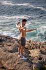 Muscular male bodybuilder with naked torso standing on seashore and doing exercises with resistance band during workout in summer — Stock Photo