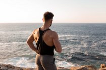Вид сзади на мускулистого спортсмена в спортивной одежде, стоящего на пляже и наслаждающегося закатом после тренировки летом — стоковое фото