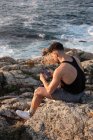 Vista lateral de alto ángulo de hombre en ropa de verano sentado en la roca cerca del mar y mensajería en las redes sociales a través del teléfono celular al atardecer - foto de stock