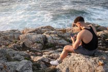Вид сбоку: мужчина в летней одежде сидит на скале у моря и смотрит в социальные сети по мобильному телефону на закате — стоковое фото