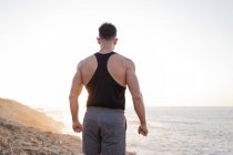 Back view of muscular male athlete in sportswear standing on beach and enjoying sundown after training in summer — Stock Photo