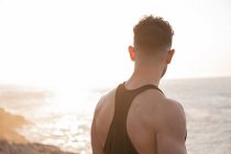 Вид ззаду м'язистого спортсмена в спортивному одязі, що стоїть на пляжі і насолоджується заходом сонця після тренувань влітку — стокове фото