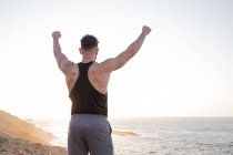 Vue arrière de l'athlète masculin musclé en tenue de sport debout sur la plage et profitant du coucher du soleil après l'entraînement en été — Photo de stock