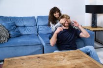 Joven mujer alegre sentada en el sofá y cubriendo los ojos del novio mientras se divierten juntos en la sala de estar - foto de stock
