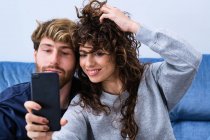 Joven barbudo hombre cerca de la mujer sonriente en ropa casual tomando selfie en el teléfono móvil - foto de stock