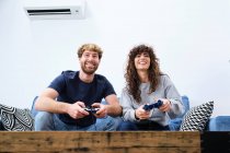 D'en bas joyeux et excité jeune couple en vêtements décontractés jouer jeu vidéo sur console dans le salon élégant — Photo de stock
