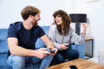 Вид сбоку веселой и взволнованной молодой пары в повседневной одежде, играющей видеоигру на консоли в стильной гостиной — стоковое фото