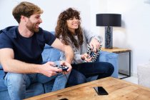 Vista lateral de pareja joven alegre y emocionada en ropa casual jugando videojuego en la consola en la elegante sala de estar - foto de stock