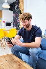 Молодой концентрированный мужчина в повседневной одежде сидит на диване и просматривает мобильный телефон в современном стильном номере — стоковое фото
