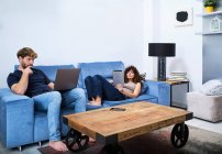 Mujer joven navegando tableta tumbado en el sofá, mientras que el hombre navegar por Internet en el ordenador portátil en apartamento moderno - foto de stock