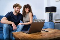 Молода щаслива пара в повсякденному одязі розмовляє онлайн на ноутбуці, сидячи на дивані у вітальні — стокове фото