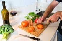 Homem anônimo de pé na cozinha enquanto corta tomates na tábua de corte com faca perto de garrafa e vinho de vidro — Fotografia de Stock