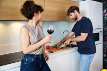Side view of young couple standing in kitchen with glass of red wine near stove and counter and cutting tomatoes on cutting board with knife near sink and microwave — Stock Photo