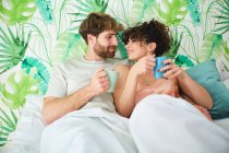 Молода пара в сплячому одязі обіймається, насолоджуючись гарячою кавою в ліжку на білій постільній білизні з подушками і ніжно дивлячись один на одного в світлій квартирі — стокове фото