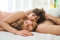 Vista lateral de la pareja joven abrazándose juntos en la cama blanca mientras usan ropa de dormir y acostados el uno sobre el otro en el dormitorio ligero - foto de stock