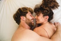 Vista superior vista lateral de la pareja joven en ropa de dormir acostada en la cama y abrazándose mientras se miran con ternura en la habitación de luz - foto de stock