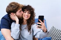 Молодий бородатий чоловік цілує усміхнену жінку в повсякденному одязі, приймаючи селфі на мобільний телефон — стокове фото