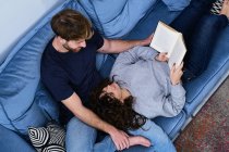 De cima jovem mulher em roupa casual deitada no sofá e livro de leitura colocando a cabeça nos joelhos do namorado — Fotografia de Stock