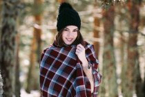 Content female wrapped in warm plaid standing in snowy woods and looking at camera — Stock Photo