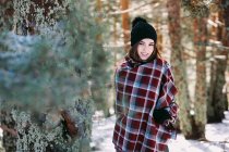Contenuto femminile avvolto in caldo plaid in piedi in boschi innevati e guardando la fotocamera — Foto stock