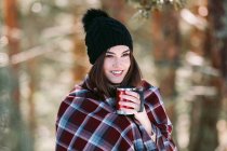 Donna avvolta in un caldo plaid seduta sul tronco d'albero nella foresta invernale innevata e che si gode una bevanda calda dalla tazza — Foto stock
