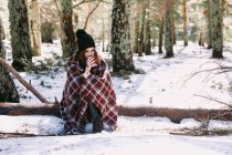 Female wrapped in warm plaid sitting on tree trunk in snowy winter forest and enjoying hot drink from cup — Stock Photo
