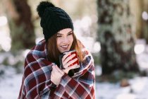 Жінка, загорнута в теплий плед, сидить на стовбурі дерева в засніженому зимовому лісі і насолоджується гарячим напоєм з чашки — стокове фото