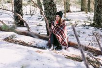 Женщина, завернутая в теплую клетку, сидит на стволе дерева в снежном зимнем лесу и наслаждается горячим напитком из чашки — стоковое фото
