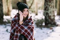 Cheerful female wrapped in warm plaid sitting on tree trunk in winter forest and talking mobile phone on sunny day — Stock Photo