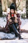 Весела жінка, загорнута в теплу плед, сидить на стовбурі дерева в зимовому лісі і переглядає мобільний телефон в сонячний день — стокове фото