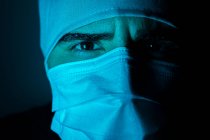 Primer plano del cirujano masculino en máscara médica mirando a la cámara en habitación oscura con luz de neón azul - foto de stock