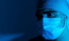 Closeup de cirurgião masculino em máscara médica olhando para longe no quarto escuro com luz de néon azul — Fotografia de Stock