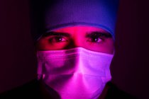 Nahaufnahme eines männlichen Chirurgen in medizinischer Maske, der in einem dunklen Raum mit blauem und rotem Neonlicht in die Kamera blickt — Stockfoto