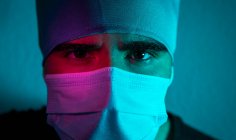 Gros plan du chirurgien masculin en masque médical regardant la caméra dans la pièce sombre avec la lumière bleue et rouge néon — Photo de stock