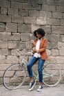 Delighted African American female standing near parked bike with takeaway drink and messaging on social media on smartphone on street — Stock Photo