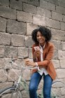 Delighted African American female standing near parked bike with takeaway drink and messaging on social media on smartphone on street — Stock Photo