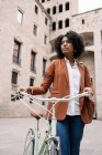 De baixo de mulher negra em estilo casual inteligente andando ao longo da rua com bicicleta e olhando para longe — Fotografia de Stock