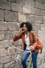 Délicieuse femme afro-américaine debout près du vélo garé avec boisson à emporter et téléphone parlant dans la rue — Photo de stock